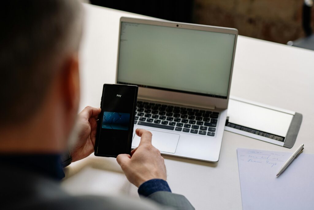 Over the shoulder view of businessman using mobile phone for digital payment beside a laptop in office.