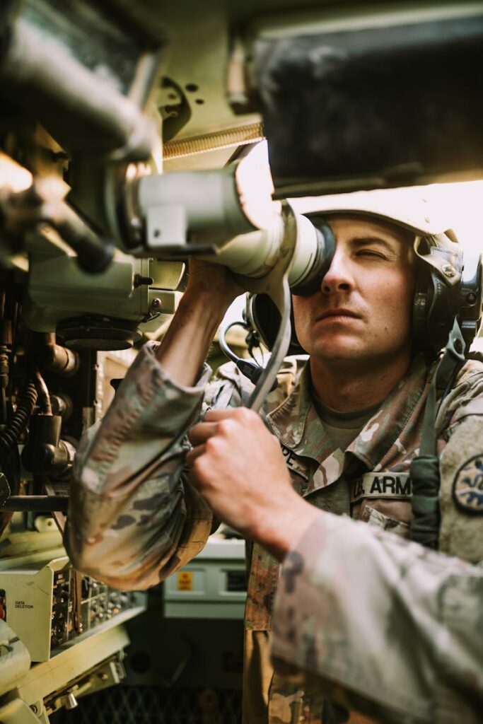 A soldier in uniform operates a periscope inside a military vehicle cockpit.