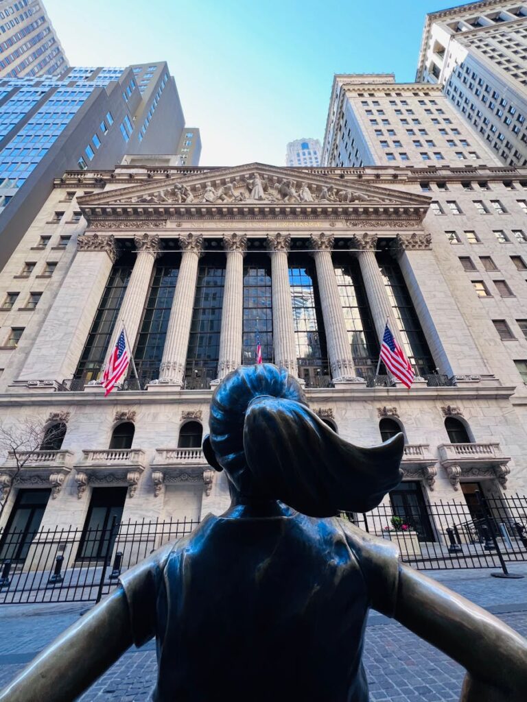 Iconic Fearless Girl Statue in front of the New York Stock Exchange building in daylight.
