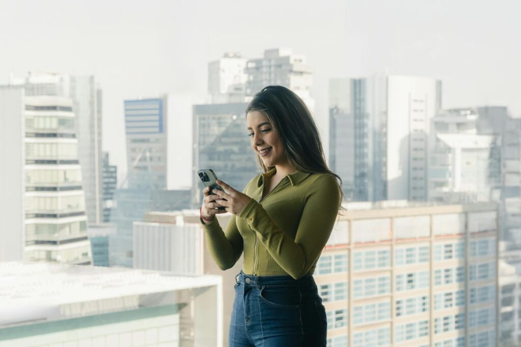 Smiling woman using phone with city skyline in the background, enjoying urban view.