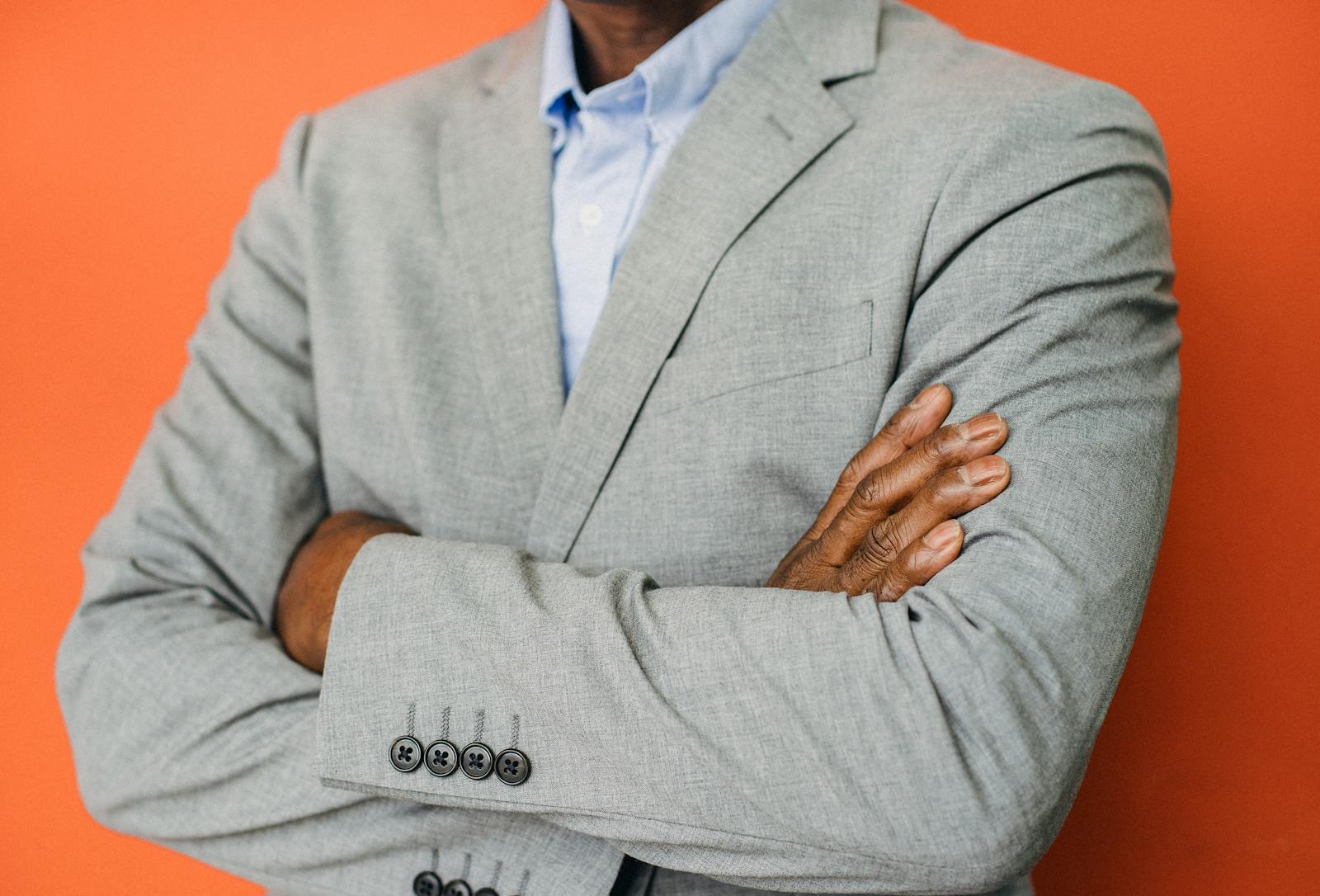Close-up of a confident businessman in a gray suit with arms crossed against a vibrant orange background.