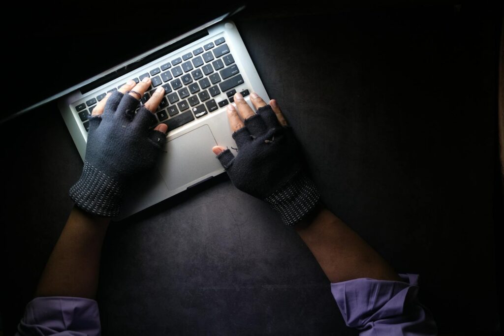 Hacker in fingerless gloves typing on laptop keyboard from above in a dark setting.