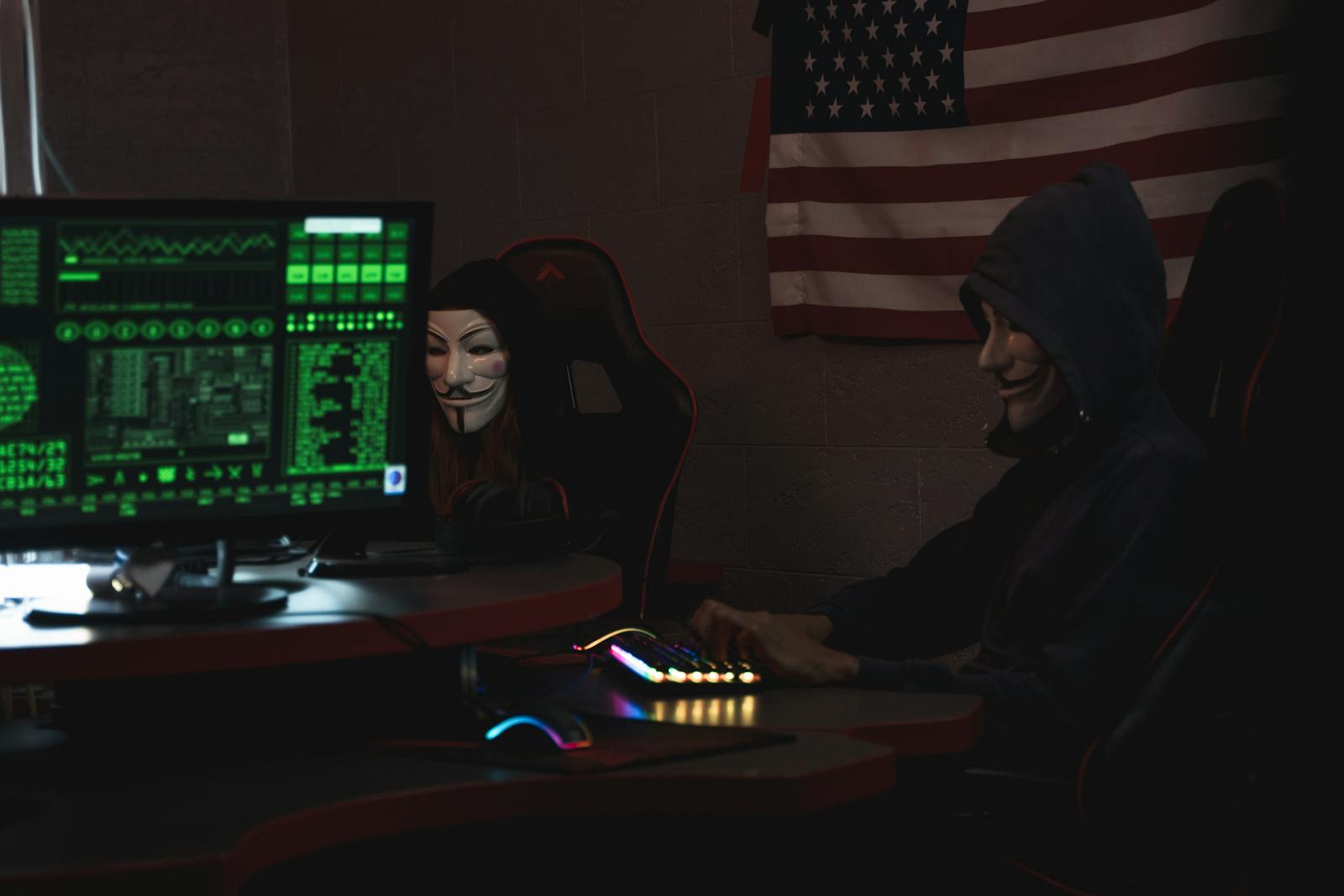 How AI Is Closing in on Human New Cybercriminals Two anonymous individuals wearing hacker masks use computers in a dark room with an American flag backdrop, highlighting cyber security themes.