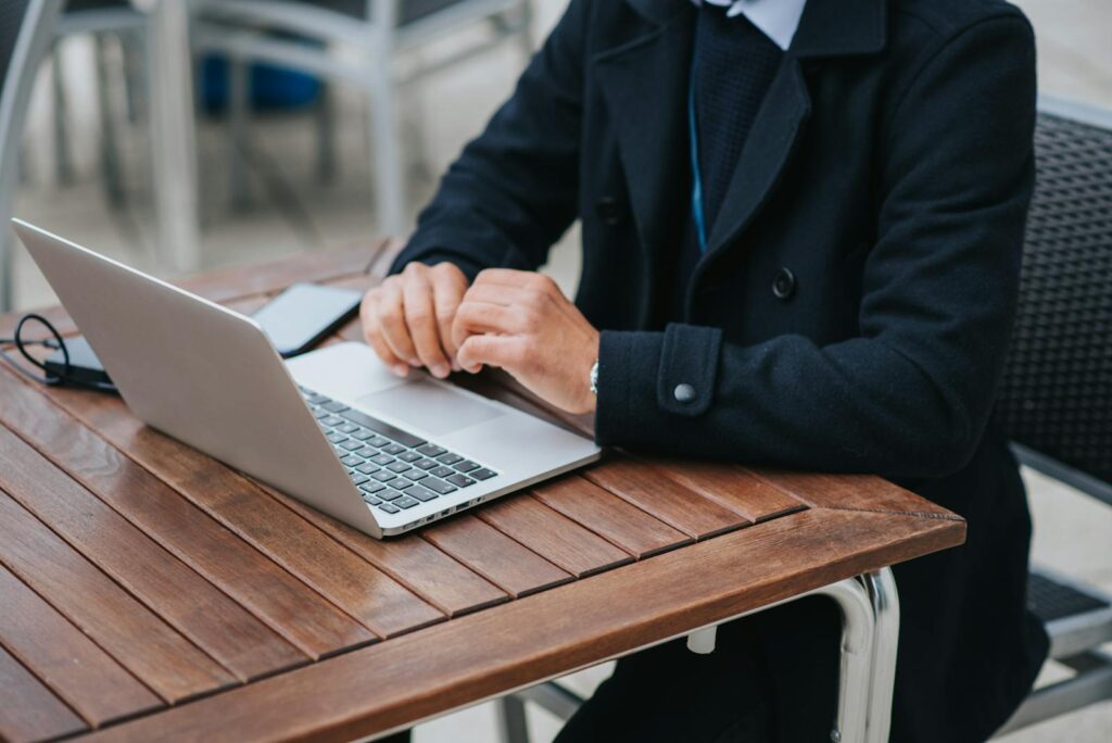 Professional man using a laptop at an outdoor café table with a smartphone, working remotely.