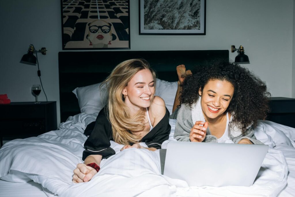 The New Weird Tech Era of 2025 on Emotional Machines What We Really Want From Technology Two young women smiling and lying on a bed, enjoying a laptop together in a cozy bedroom setting.