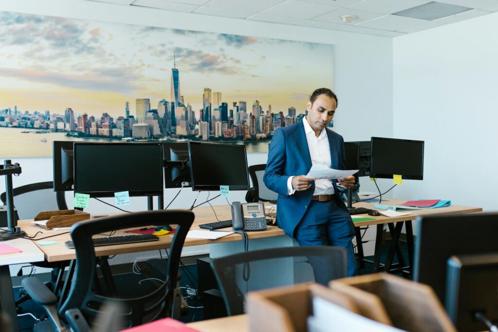 A man in a suit reviews documents in a modern office setting with computers and skyline art.