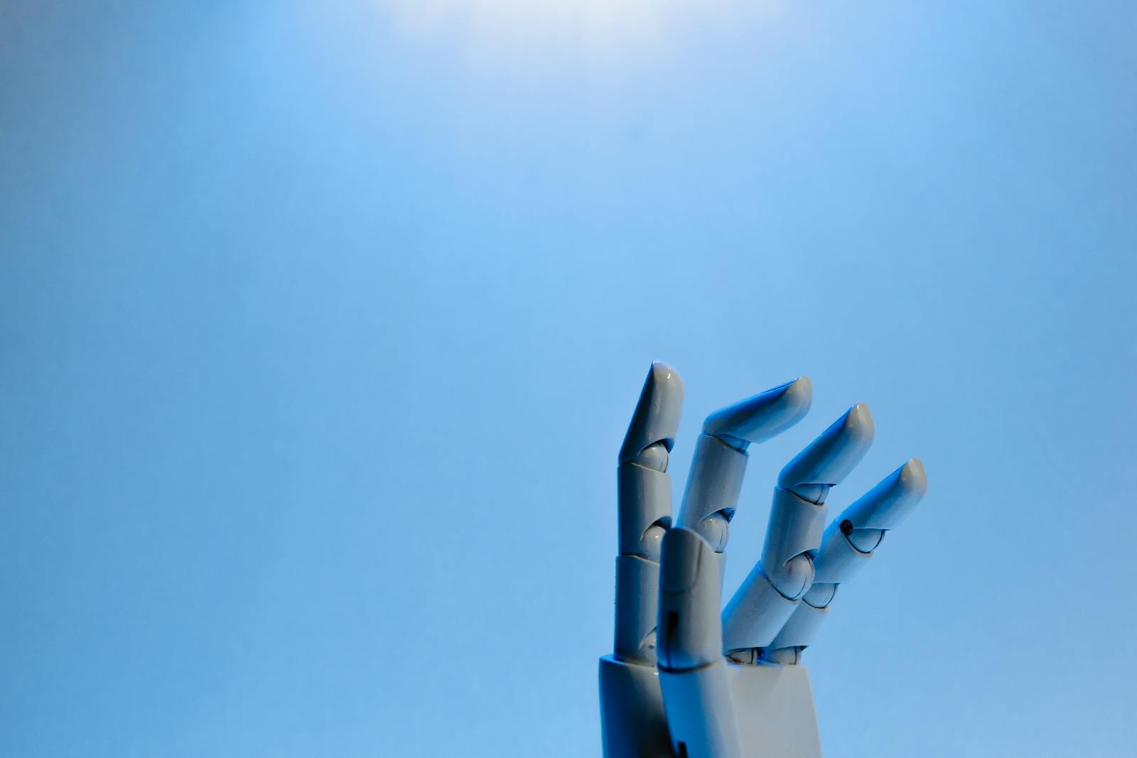 Why the New AI Boom’s Risks Are Being Passed Around Robotic hand with articulated fingers reaching towards the sky on a blue background.