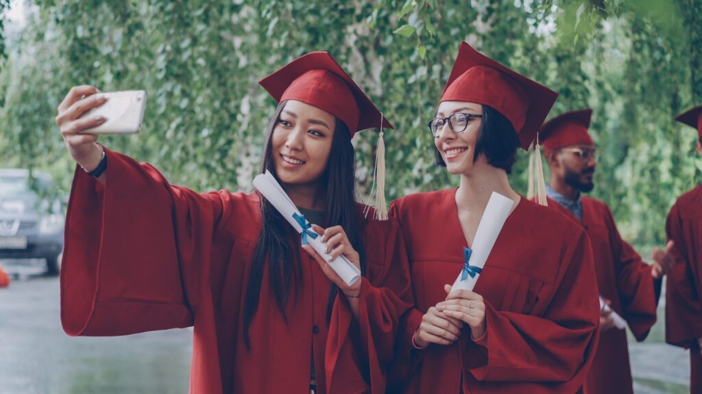 Two graduates in red gowns taking a selfie with diplomas.