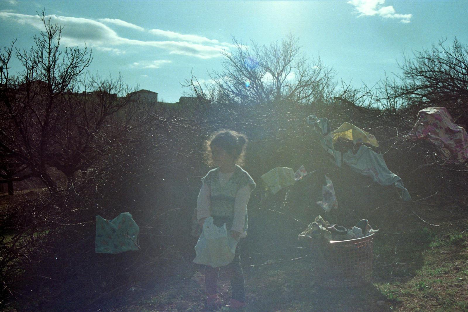 A young child stands in the sunny outdoors of Hekimhan, Türkiye, amidst nature.