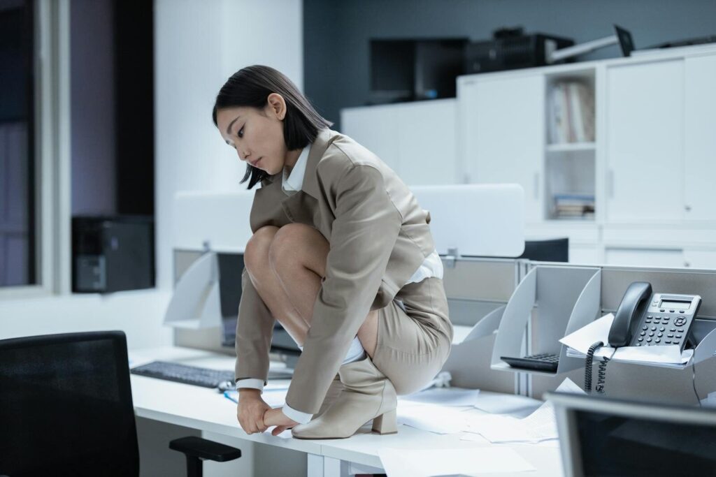 AI Anxiety Is Spreading And Could Spark New Workers’ Movement Asian businesswoman crouching on a desk in an office, depicting stress and overwork.
