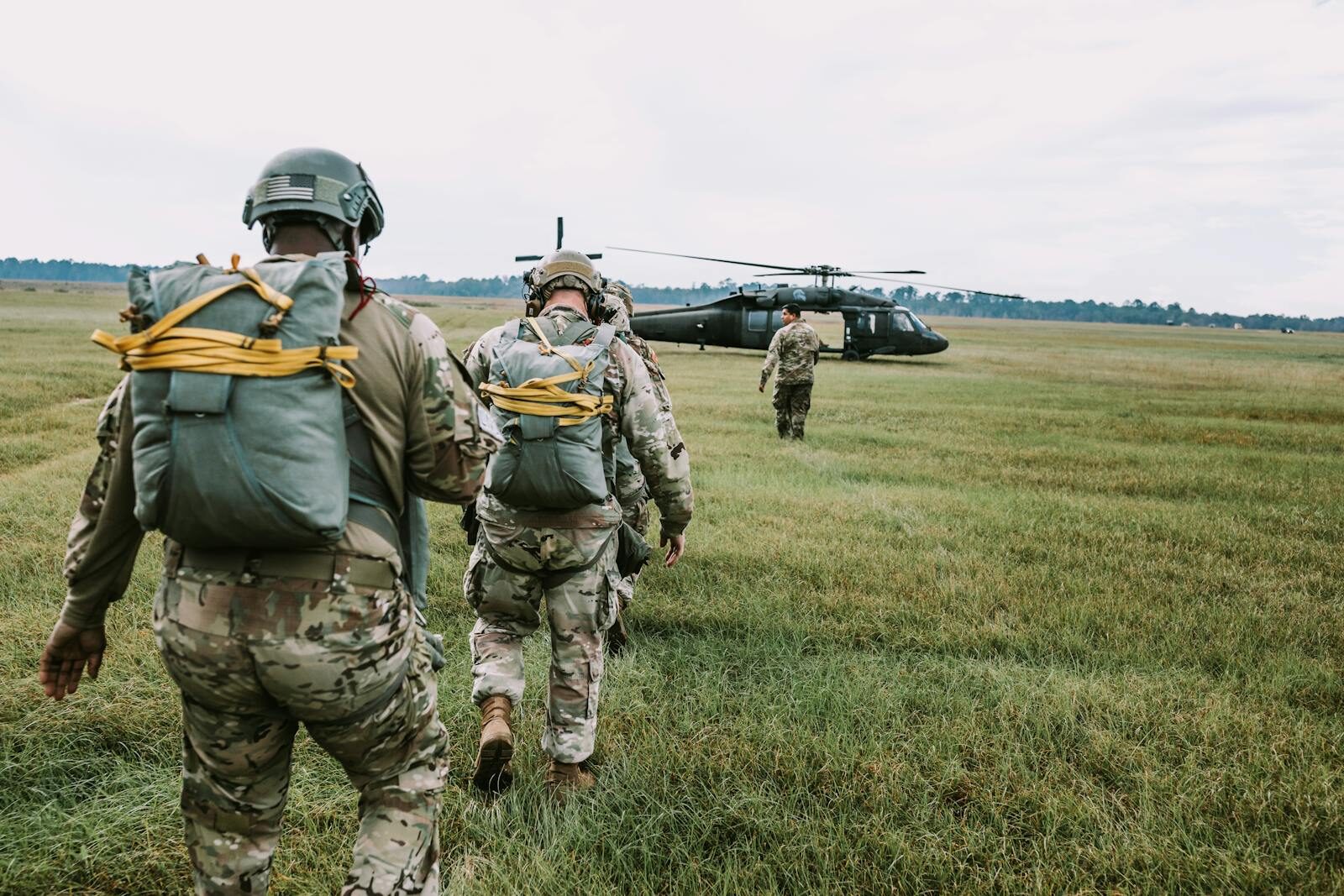 Military personnel in uniforms and gear walk toward a helicopter on a grassy field.