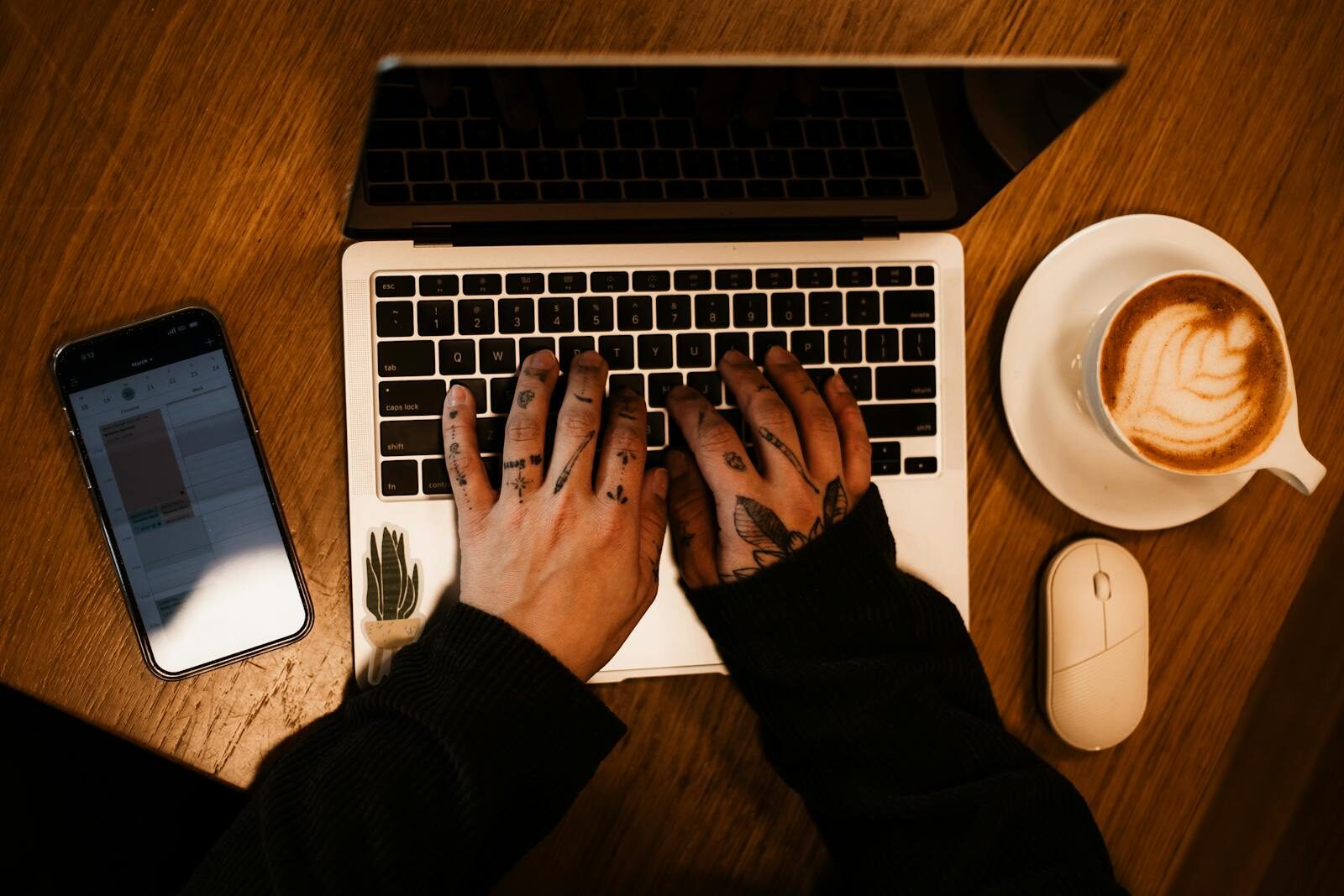 Tattooed hands on a laptop with coffee and phone, perfect workspace inspiration.