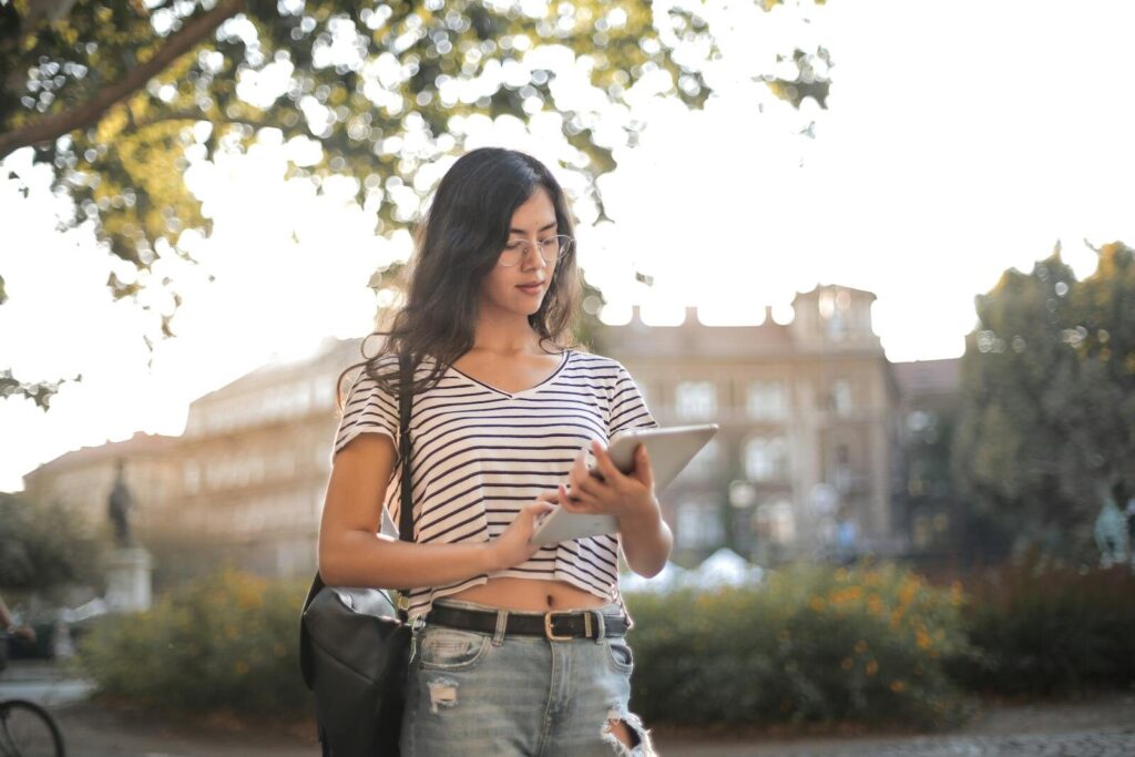 A young woman in a striped shirt and ripped jeans using a tablet in a sunny park.