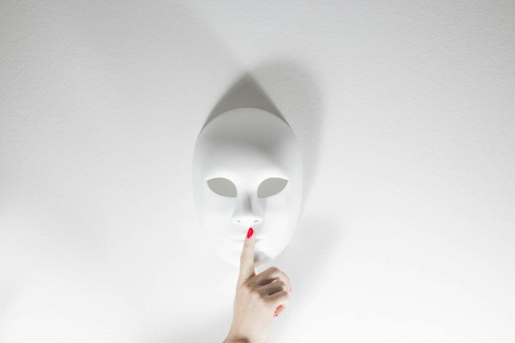 A white mask with a human hand gesturing silence on a bright white background, creating a mysterious vibe.