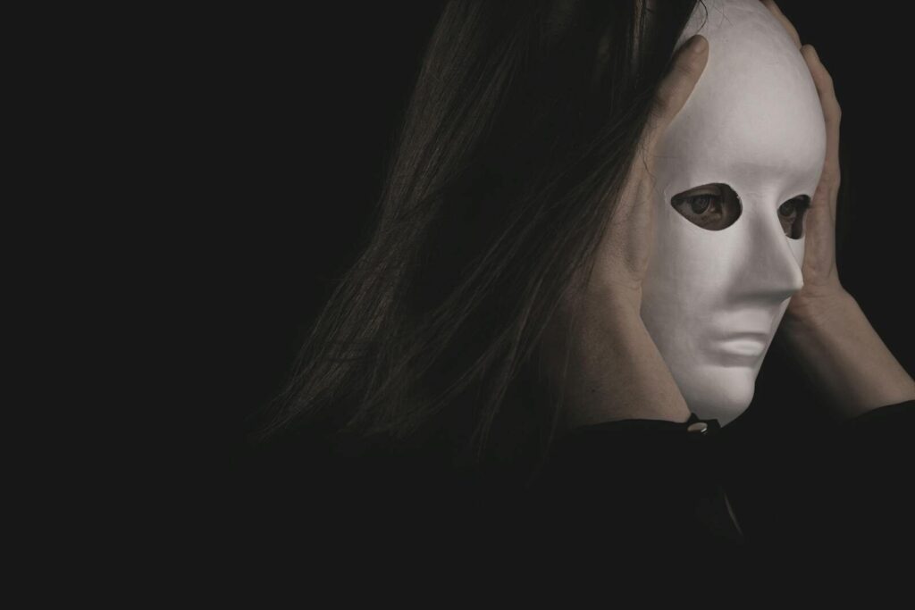 A person wearing a white mask held with both hands against a dark background, creating a mysterious atmosphere.