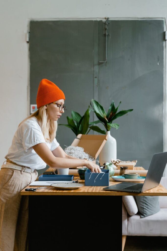 A young woman in a beanie packs boxes at a stylish wooden table in a home office.