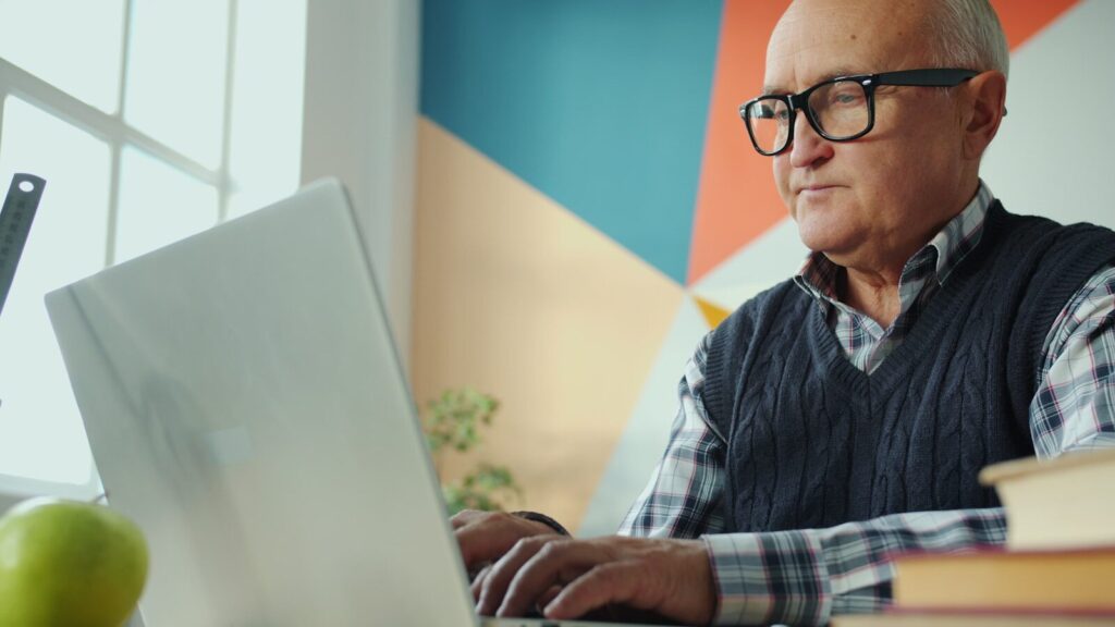 Elderly man with glasses working on a laptop.