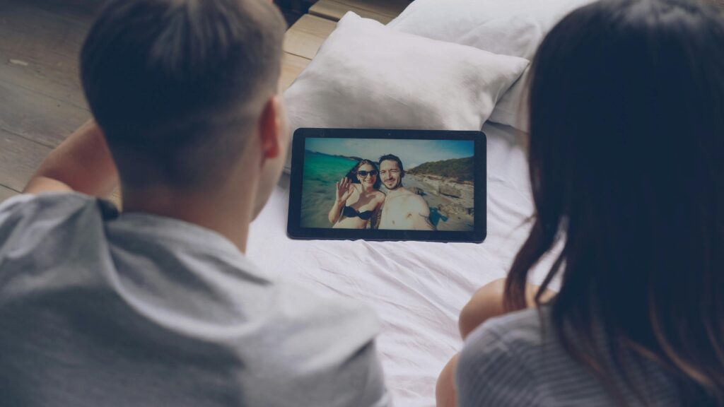 A couple watches vacation photos on a tablet while sitting on their bed.