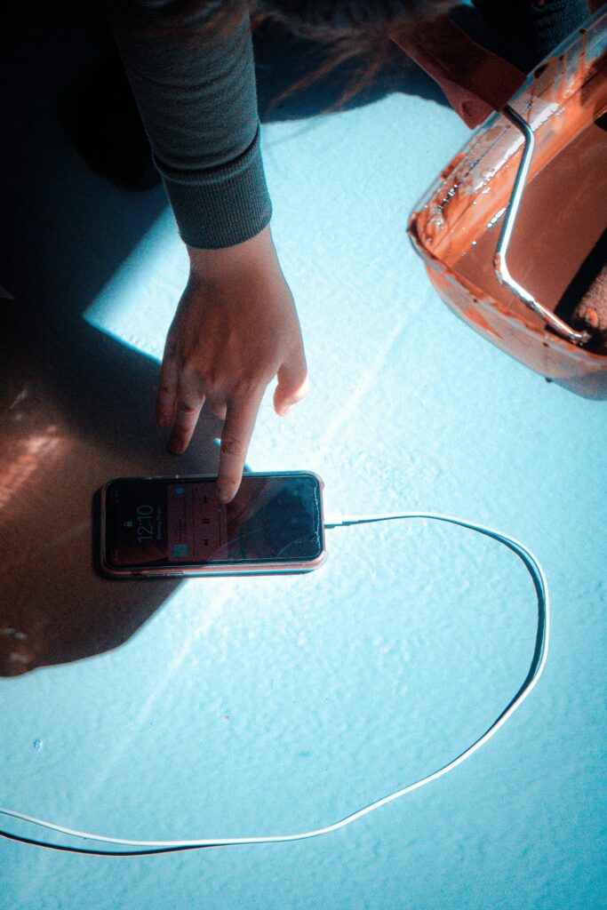 A person touches a smartphone connected to a charger next to a paint roller in a brightly lit room.