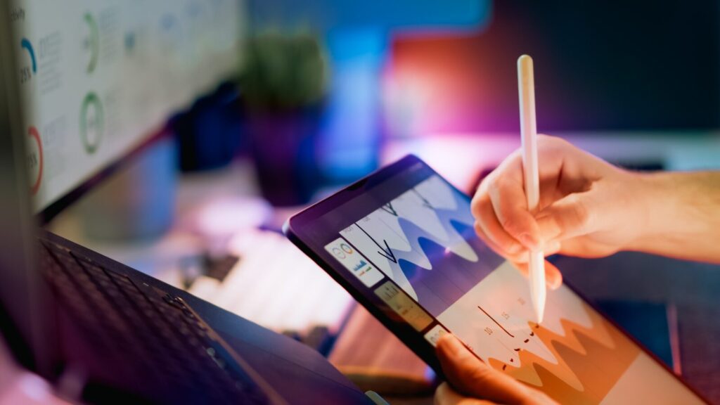 AI Is the Biggest New Tech “Tsunami” Ever, Says John Doerr Person using stylus on tablet with charts.