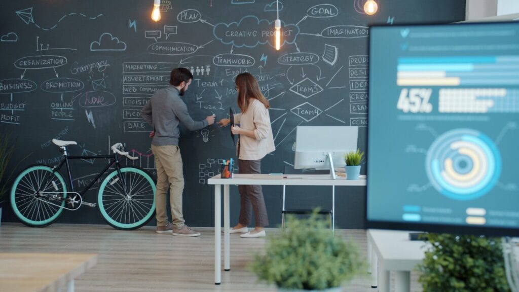 Inside Rise of New “Tokenmaxxing” and Why Companies Use More AI Two people collaborating on a chalkboard with a bicycle.