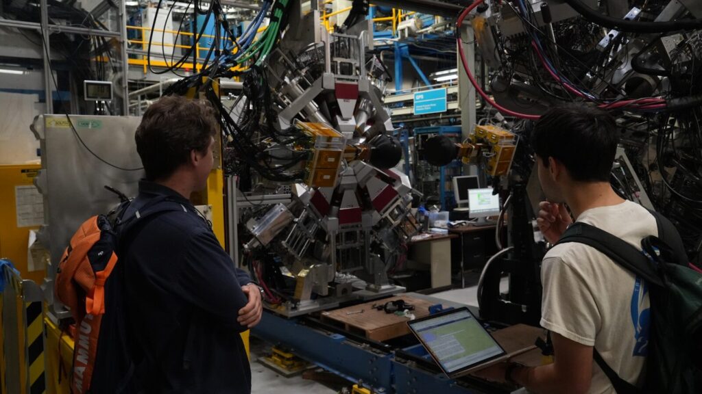Two men observe complex scientific machinery in a laboratory.